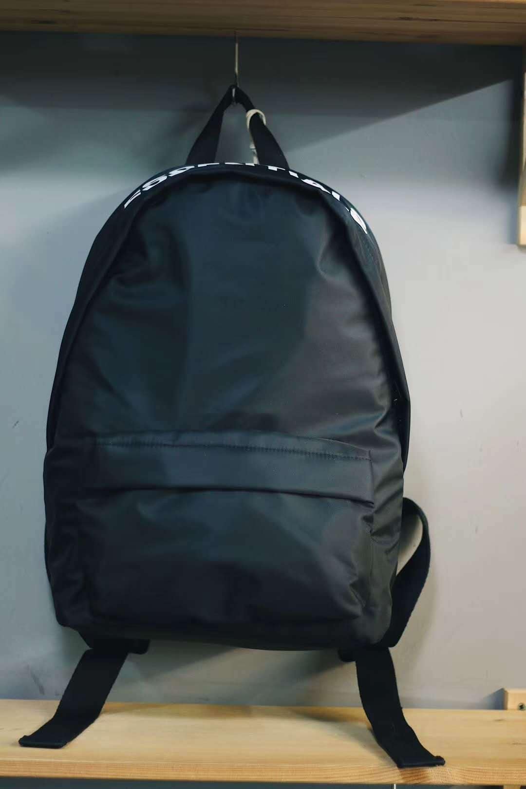 Black backpack with white text at the top, placed on a wooden shelf, perfect for carrying K-pop merchandise and accessories.