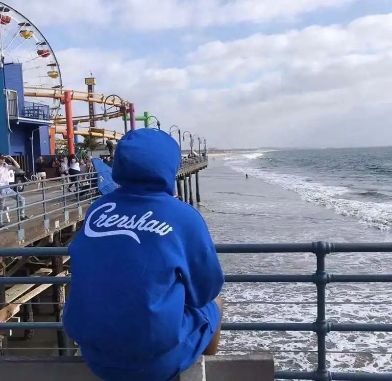 -Close-up of a person wearing a blue hoodie with "Creshwar" printed on the back, sitting by the seaside pier with amusement park rides in the background.