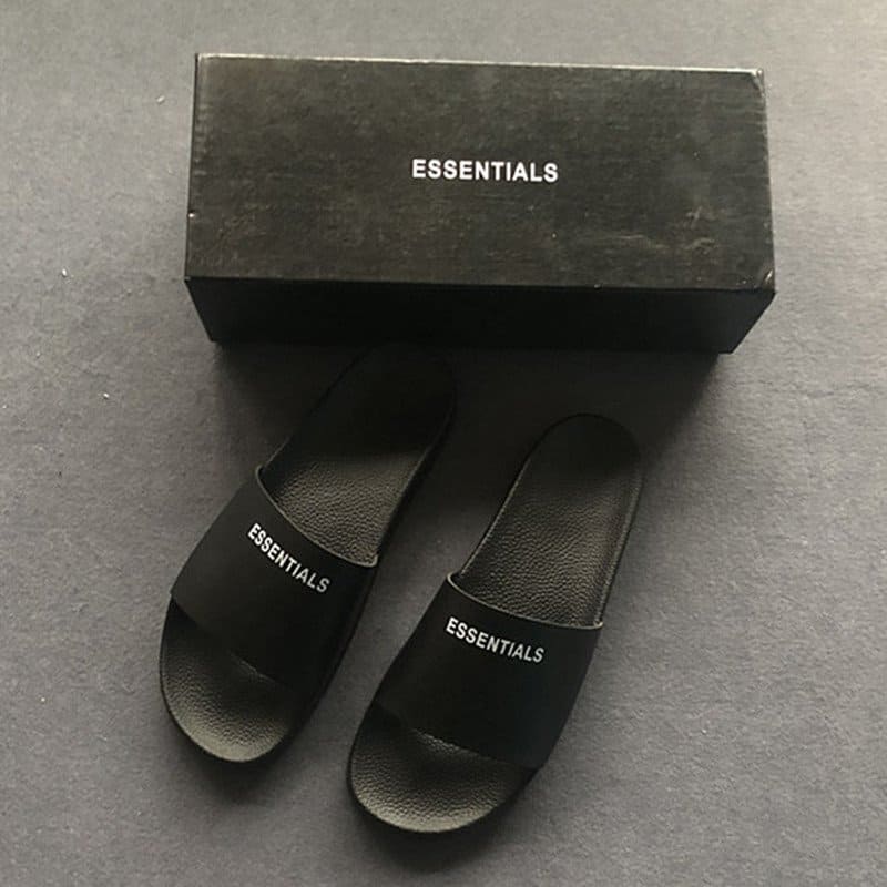 Black slide sandals with "ESSENTIALS" logo, superb for casual wear, minimalistic design, perfect for K-pop fans, comfortable and stylish footwear option for everyday use.