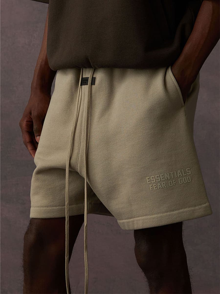 Minimalist beige shorts with "ESSENTIALS FEAR OF GOD" embroidered on the side, styled with a dark top; contemporary fashion style, unisex streetwear.