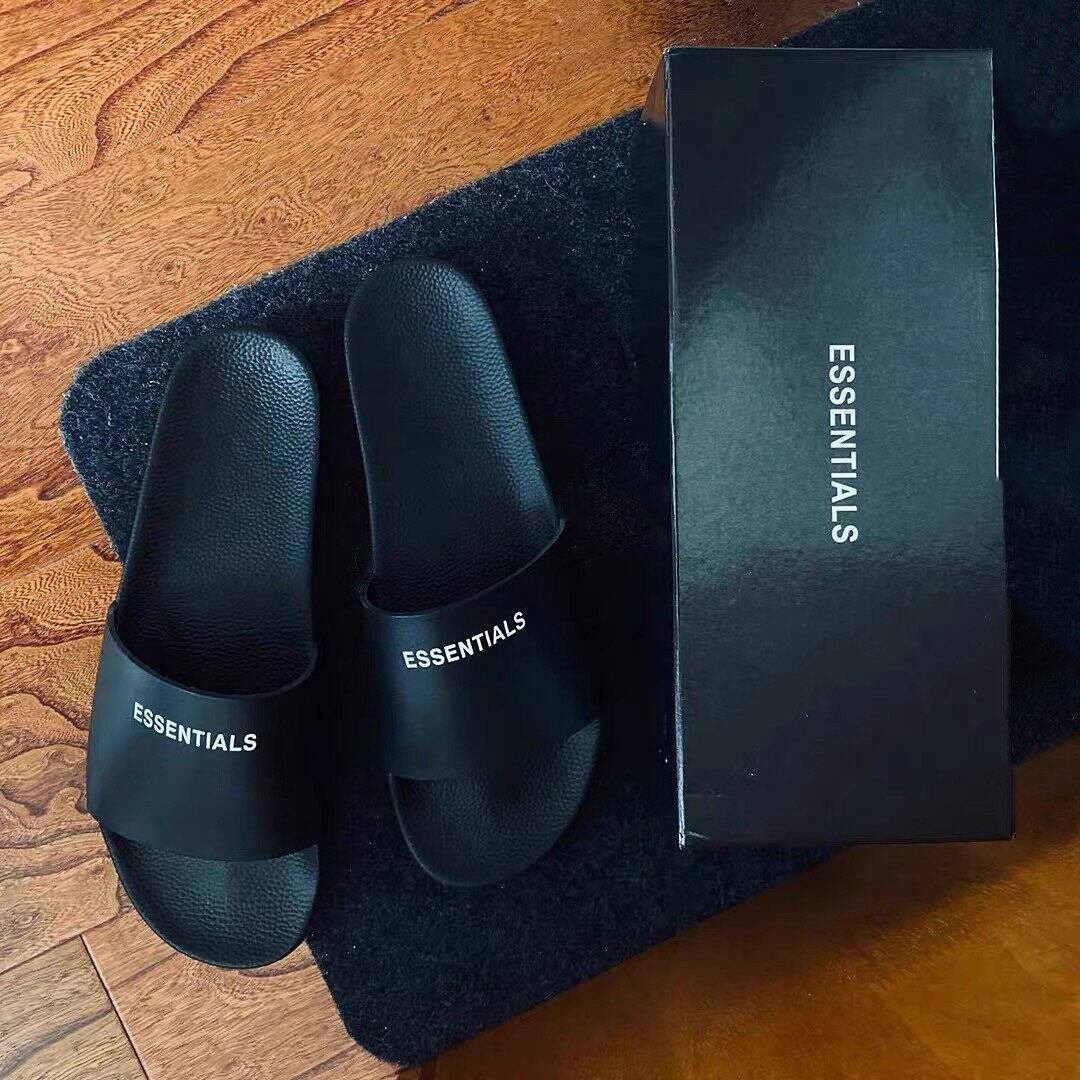Black essentials slides with logo, on a black mat, next to their box, minimalistic and stylish design.
